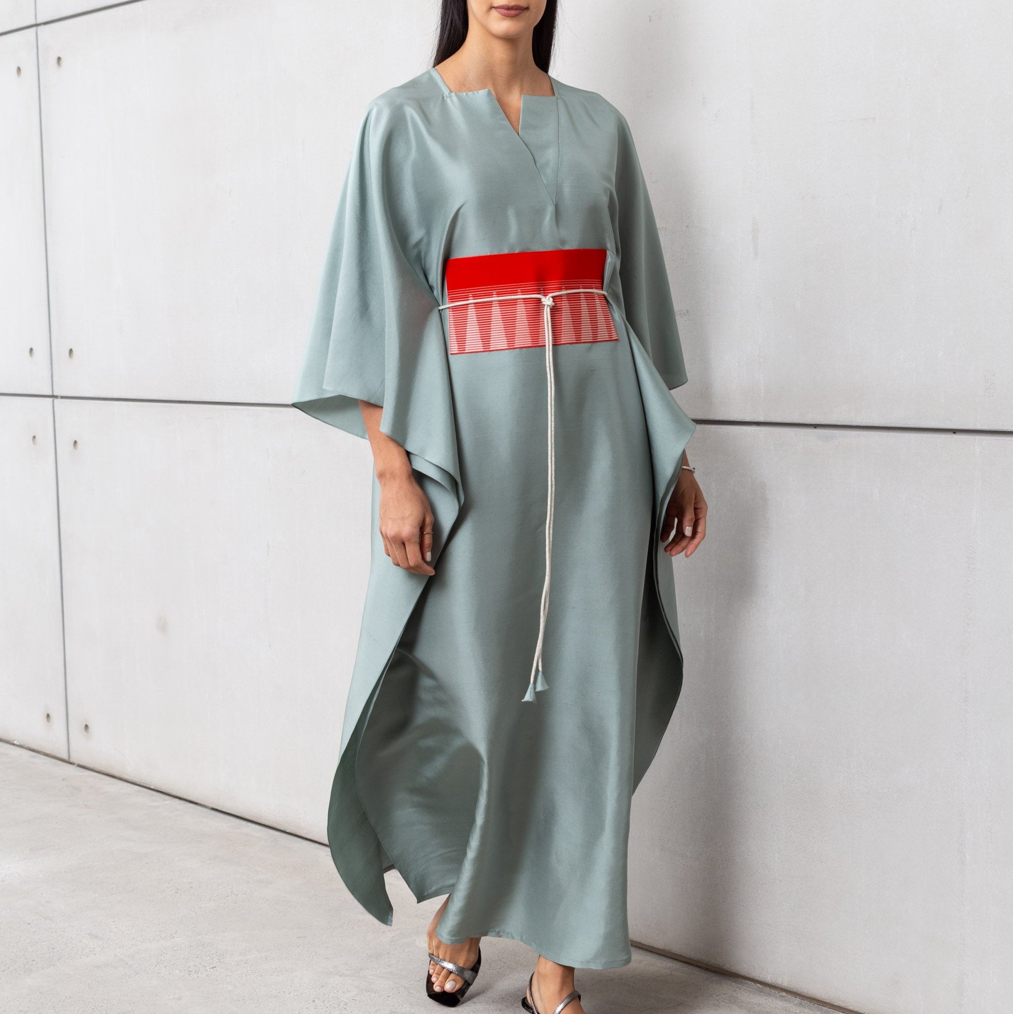 A woman wearing a pistachio-colored luxury silk kaftan with a red belt, standing against a gray backdrop.