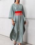 A woman wearing a pistachio-colored luxury silk kaftan with a red belt, standing against a gray backdrop.