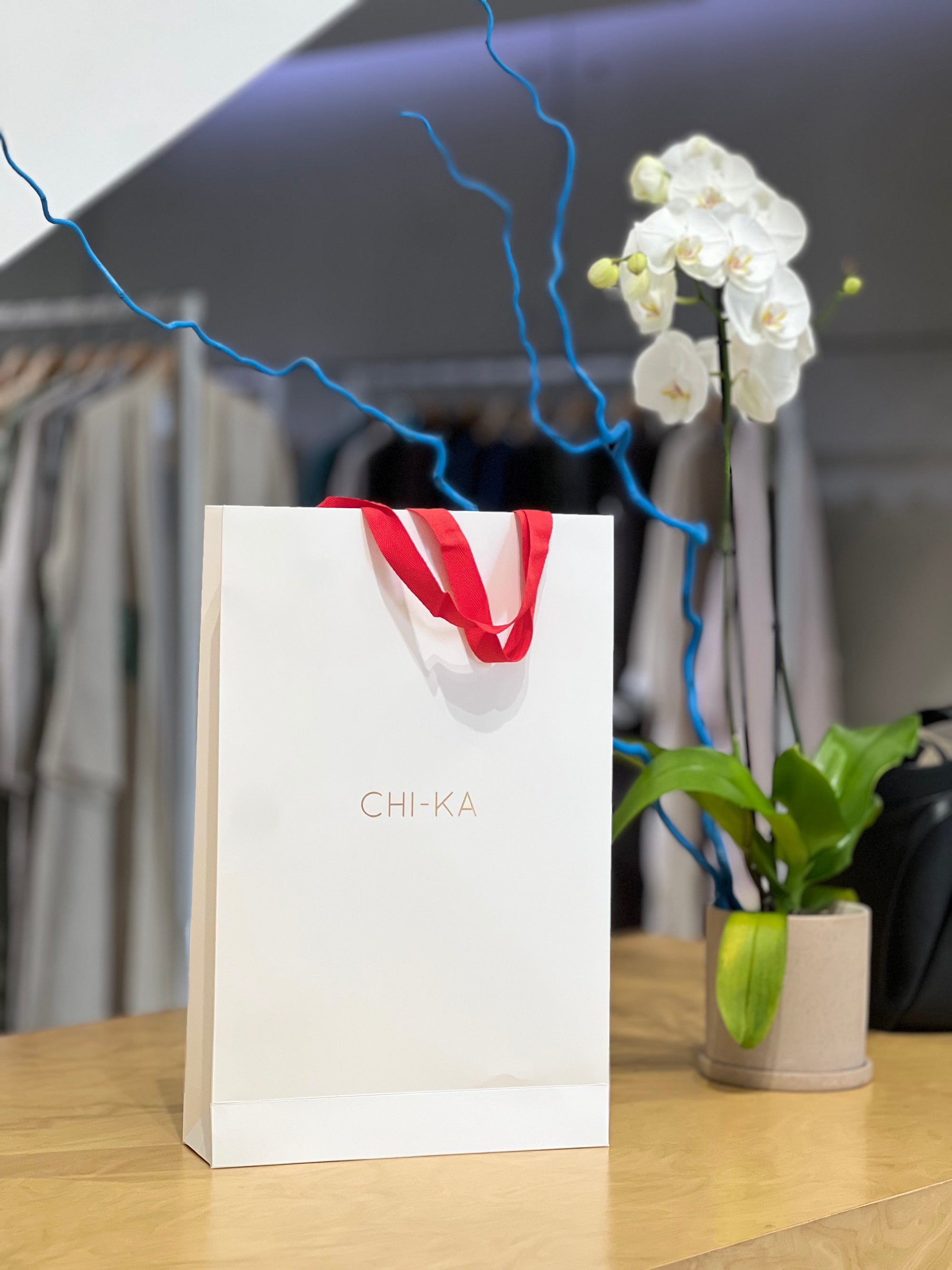 White gift bag with red ribbon and 'CHIKA' branding on a wooden surface with a blurred background.